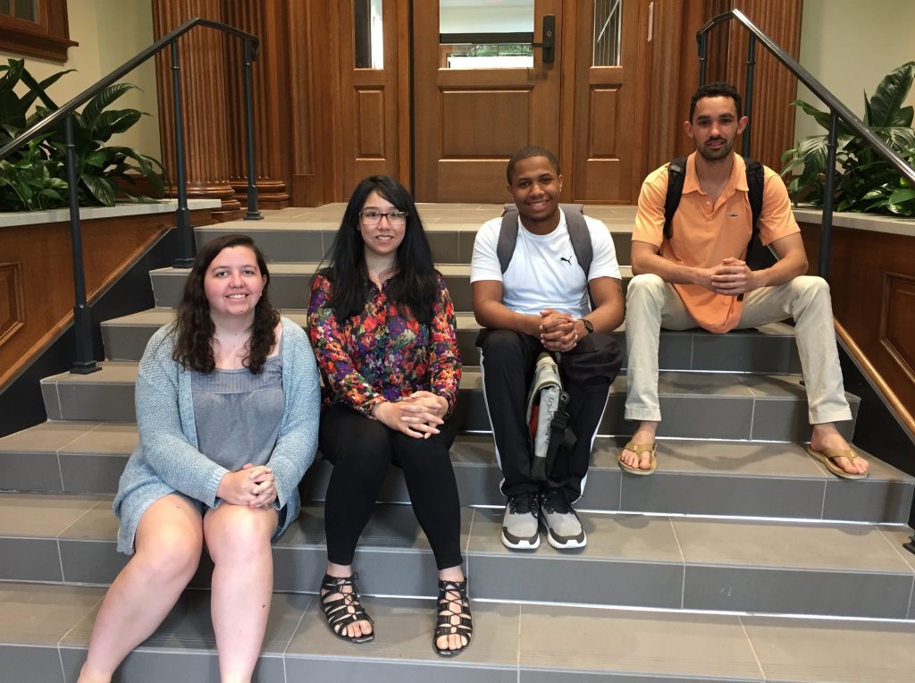 Four students smiling and sitting on steps inside.